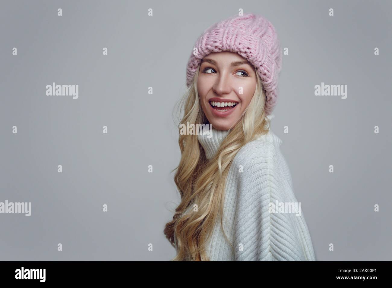 Happy young woman with long blond hair in pink knitted winter hat looking back over her shoulder with a beaming smile against a grey background Stock Photo