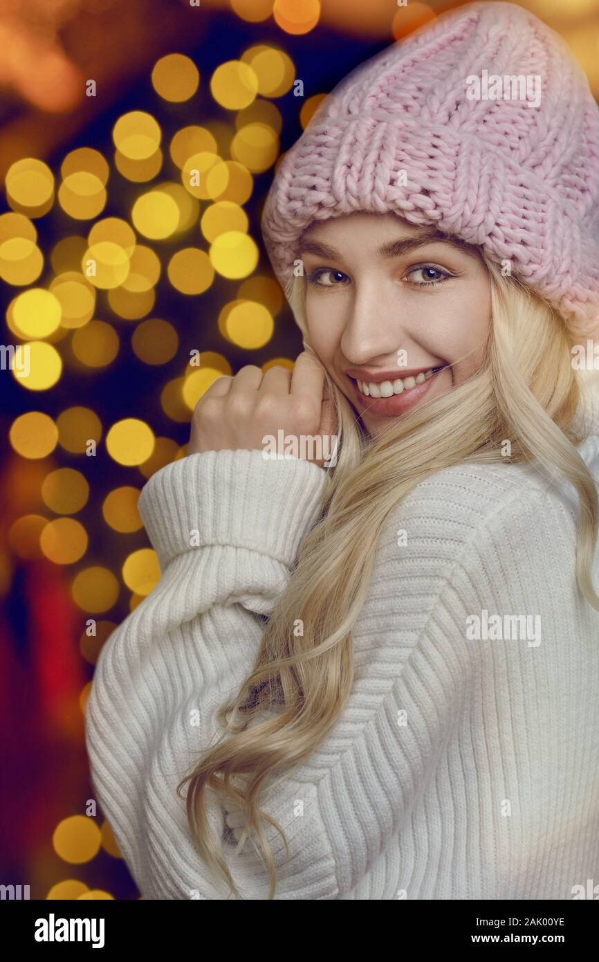 Happy young woman with long blond hair in pink knitted winter hat looking at the camera with lights in the background Stock Photo