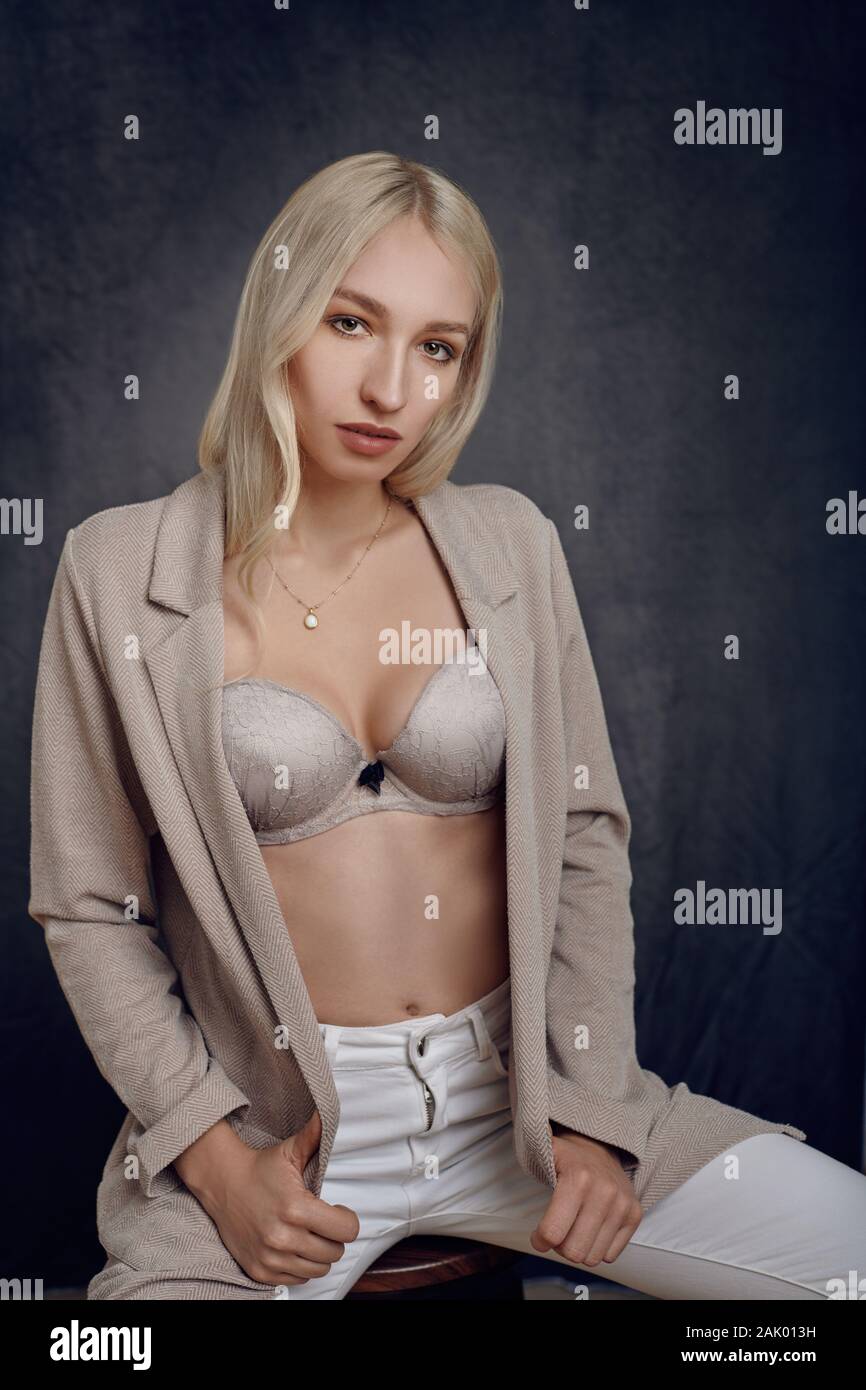 Sexy young woman with her jacket unbuttoned in a bra as she sits on a stool looking thoughtfully at the camera with a serious expression Stock Photo