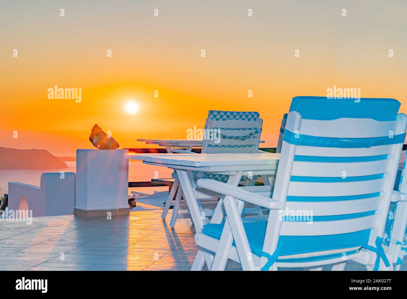 Santorini sunset from outdoors patio with outdoor furniture in white and blue under golden sky as sun sets. Stock Photo
