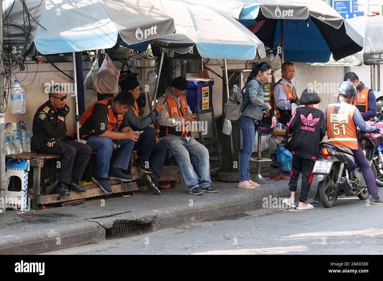 Bangkok, Thailand - December 26, 2019: Motorbike taxi drivers sitting on bench and watching their phones while waiting next customers. Stock Photo