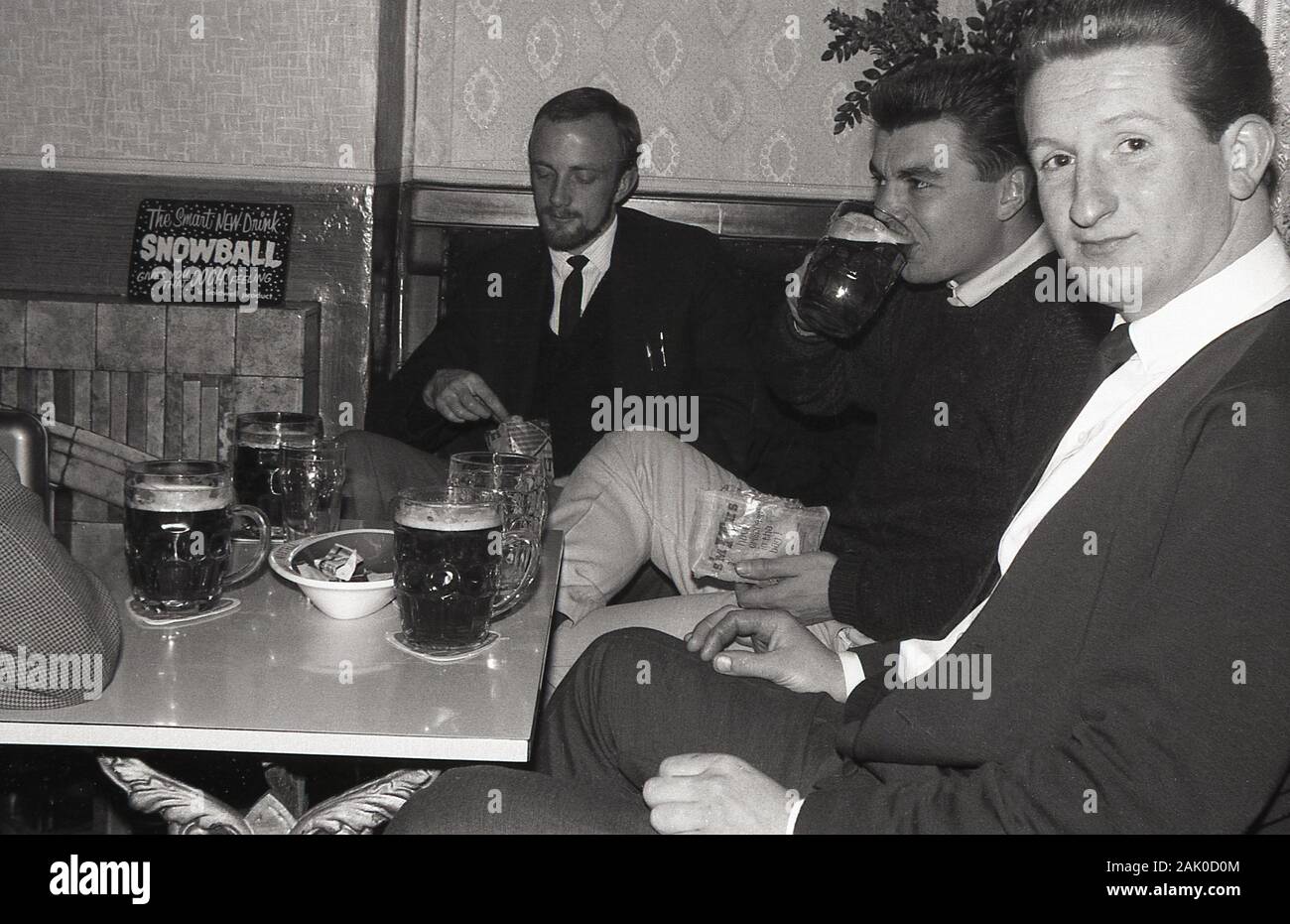 1960s, historical, three men enjoying glass tankards of beer and potato crisps sitting at a table in a corner of a bar or pub, England, UK. An advert for the new drink 'Snowball' is on the fireplace mantelpiece. Stock Photo