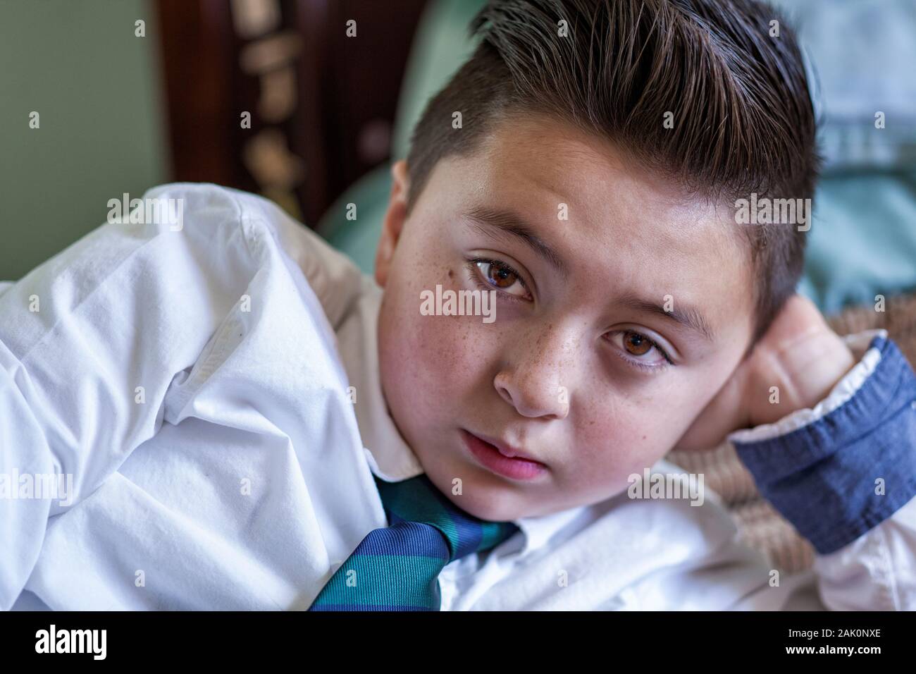 Mixed Race Asian Caucasian boy with brown Pompadour hair in white dress shirt school uniform with green and blue necktie reclining on his elbow Stock Photo
