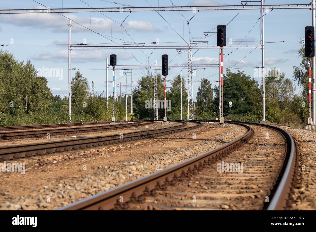 railway - train tracks with traffic lights and wires with power lines, countryside, green trees and blue sky with white clouds in the background Stock Photo