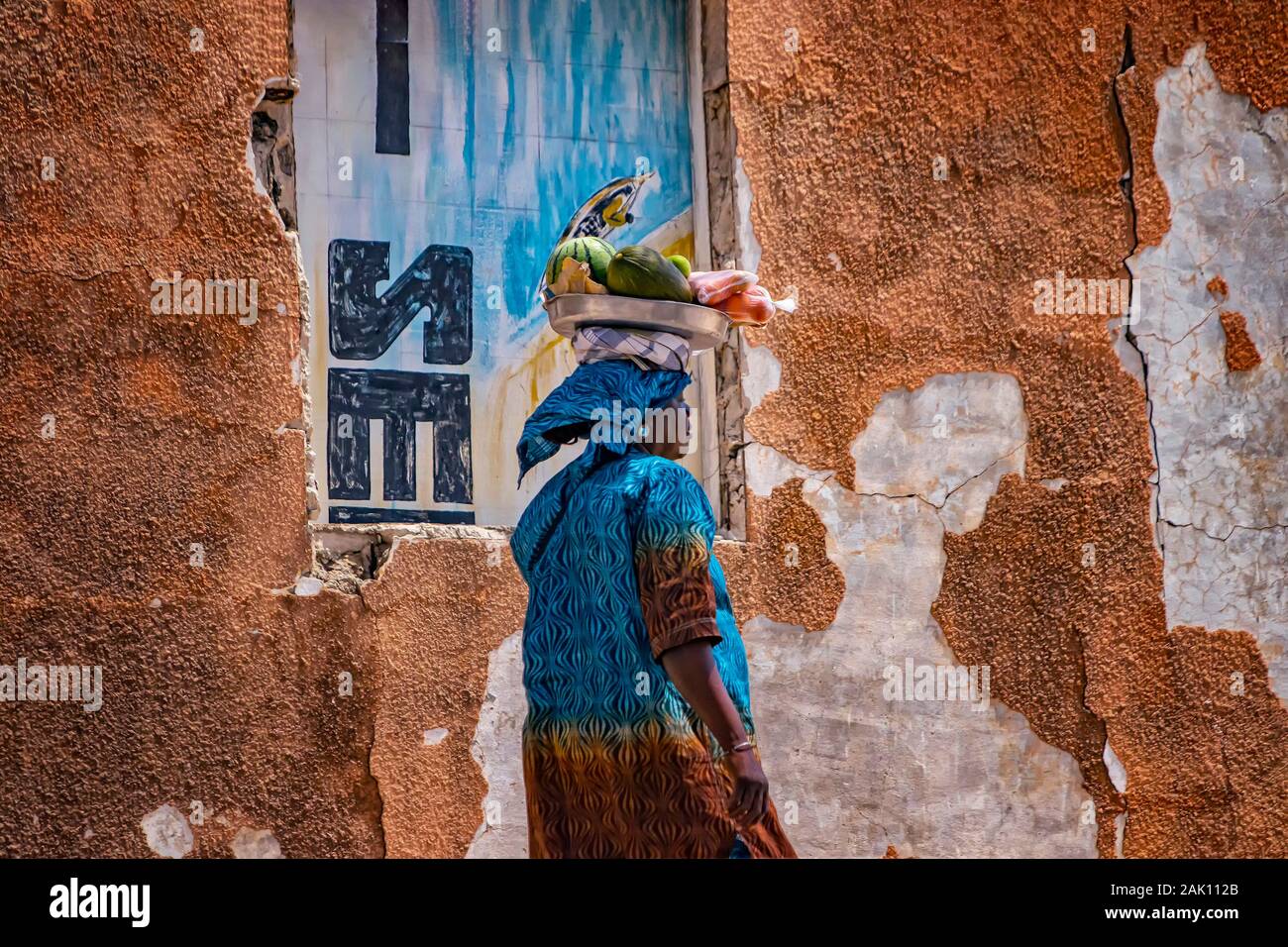 Saloum, Senegal, APRIL 26, 2019: Unidentifed Senegal woman carrying a watermelon and other fruits on her head in the city of Saloum and wearing the Stock Photo