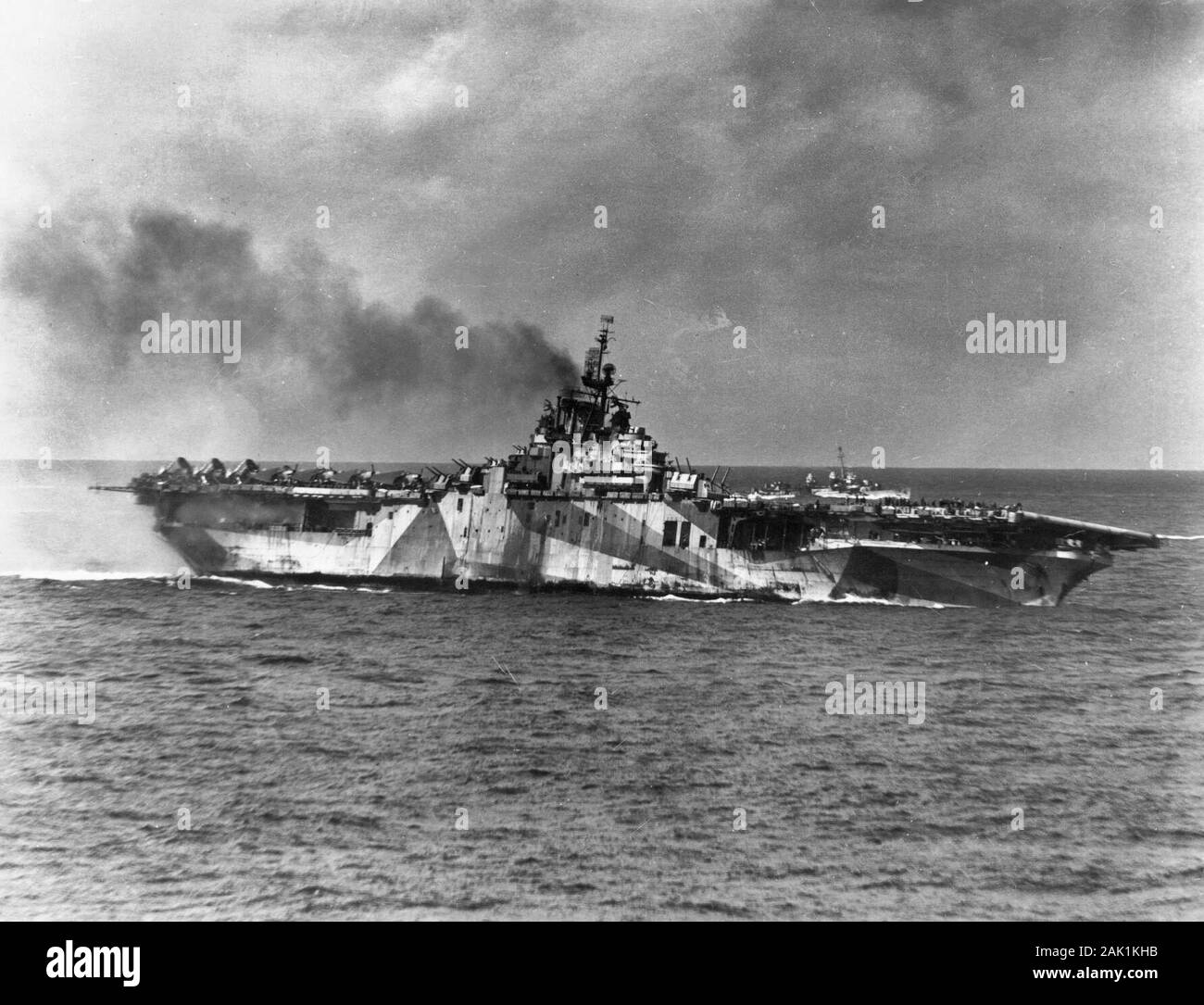 The U.S. Navy aircraft carrier USS Ticonderoga (CV-14) lists to port in the aftermath of a kamikaze attack in which four suicide planes hit the ship, 21 January 1945. Note her camouflage scheme measure 33/10A and the Fletcher-class destroyer in the background. Stock Photo