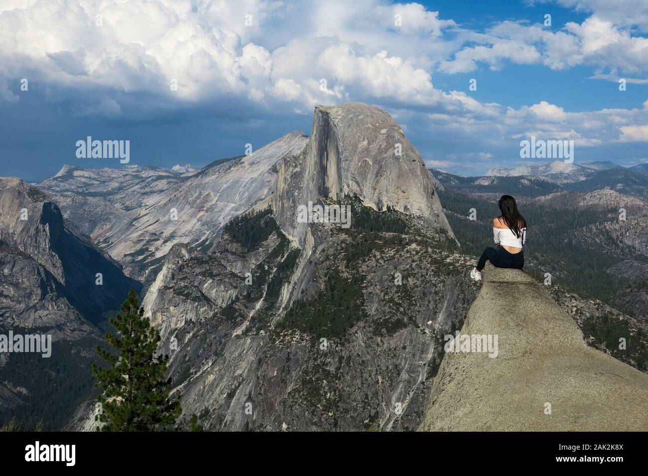 Woman hiker sitting on a dangerous rock looking out to Half Dome - California Stock Photo