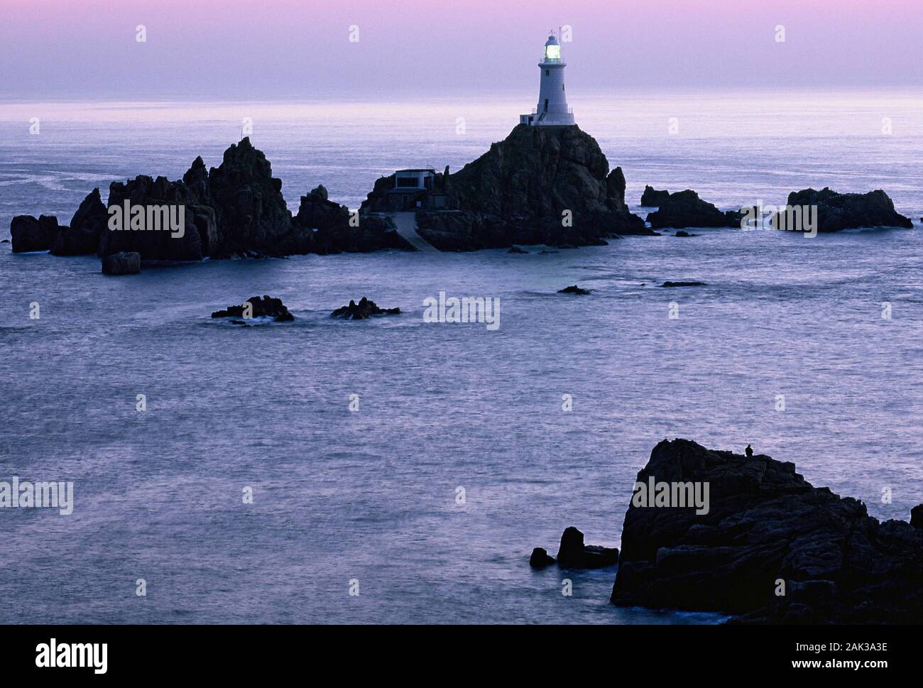 Shaded in violet colors is the Corbiere Lighthouse near La Pulente in the west of the island of Jersey at sunset. The lighthouse is situated on a rock Stock Photo