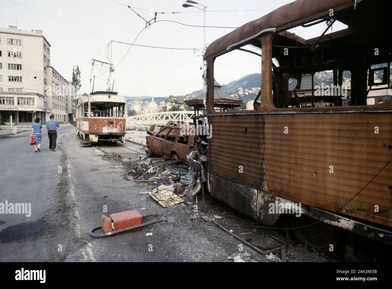 17th August 1993 During the Siege of Sarajevo:a couple walk past wrecked trams along Obala Kulina Bana during one of the frequent, though brief, ceasfires. Stock Photo