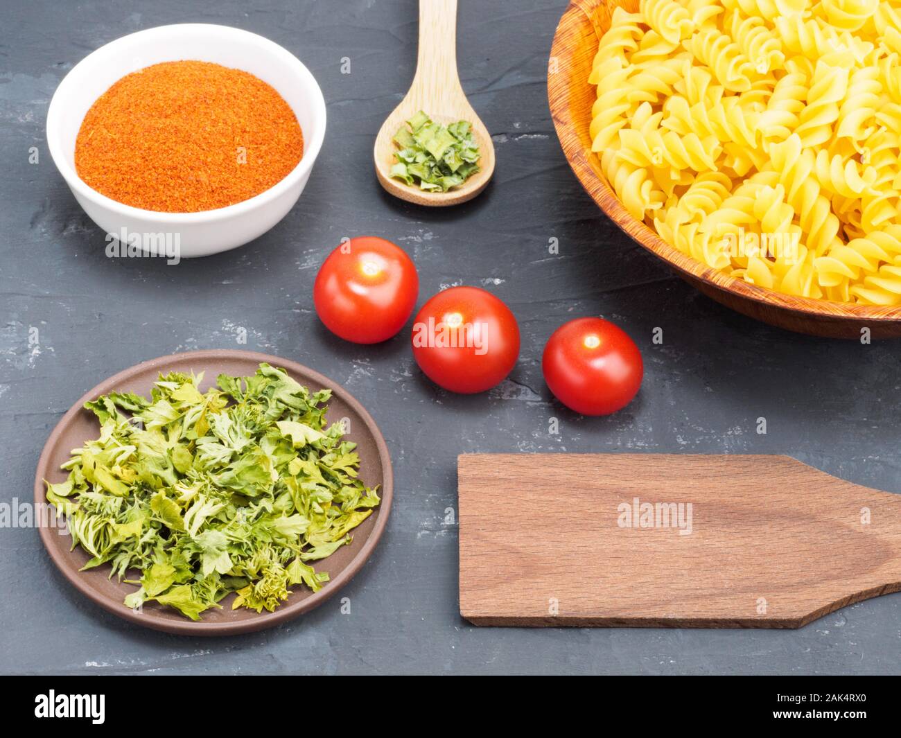 Pasta in wooden bowl, small tomatoes, red chili, green grass on a black concrete background. Healthy eating concept Stock Photo