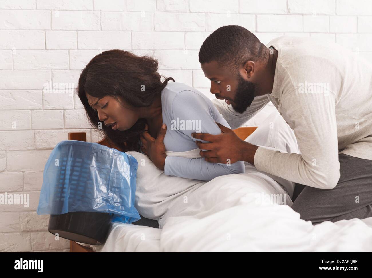 Morning sickness. Young pregnant african woman sitting on bed, holding trash, feeling nauseous during pregnancy, her man comforting her Stock Photo
