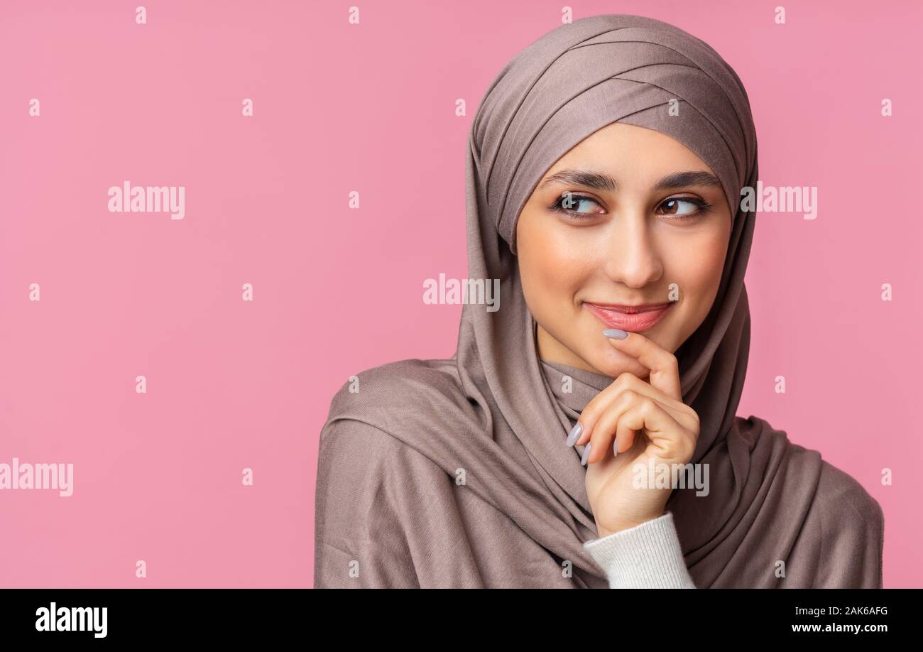 Closeup portrait of beautiful smiling muslim woman in hijab smirking, thinking about something and looking aside at copy space on pink background. Stock Photo