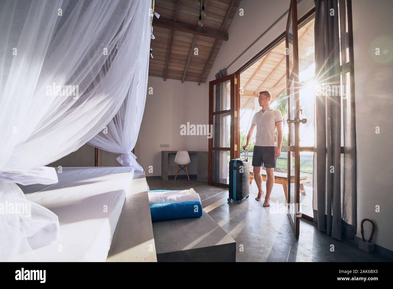 Luxury accommodation in tropical destination. Young man with suitcase entering to modern bungalow. Stock Photo