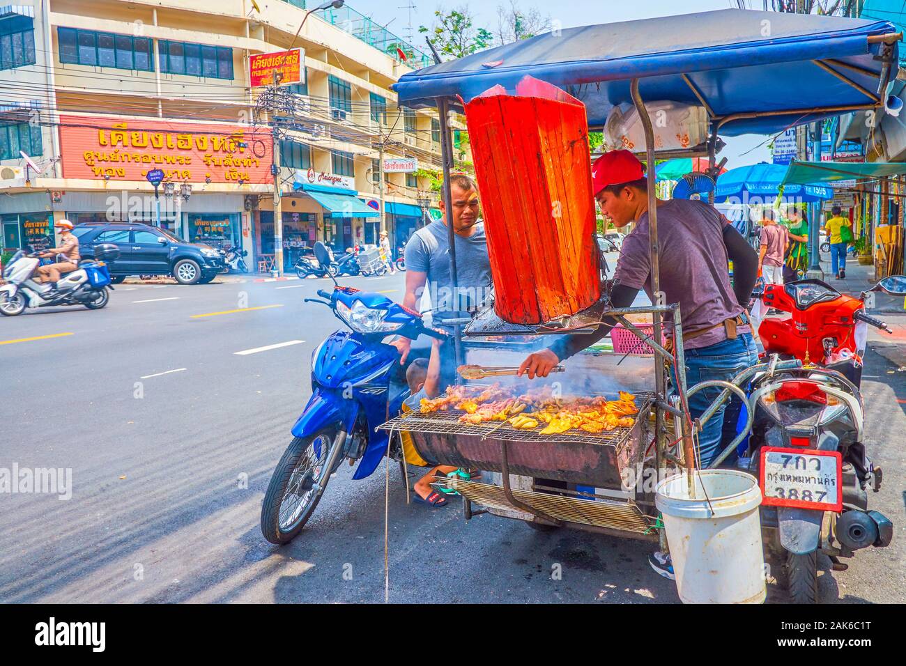 BANGKOK, THAILAND - APRIL 15, 2019: The young cook prepares street food snacks on the hand-made grill, mounted on the bike in old town, on April 15 in Stock Photo