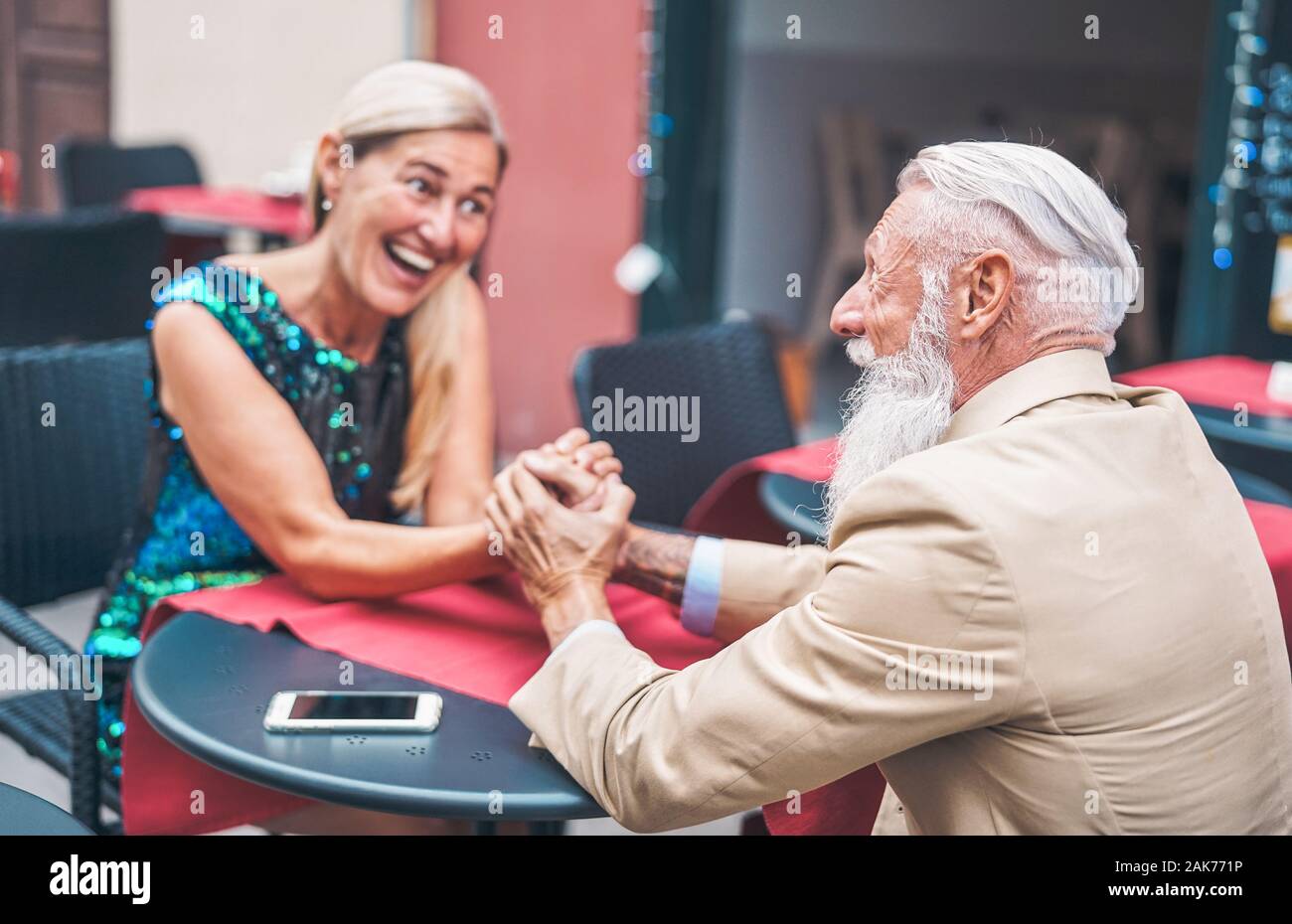 Happy senior couple holding hands while sitting in a bar outdoor - Elegant mature husband and wife looking each other while relaxing in a restaurant Stock Photo