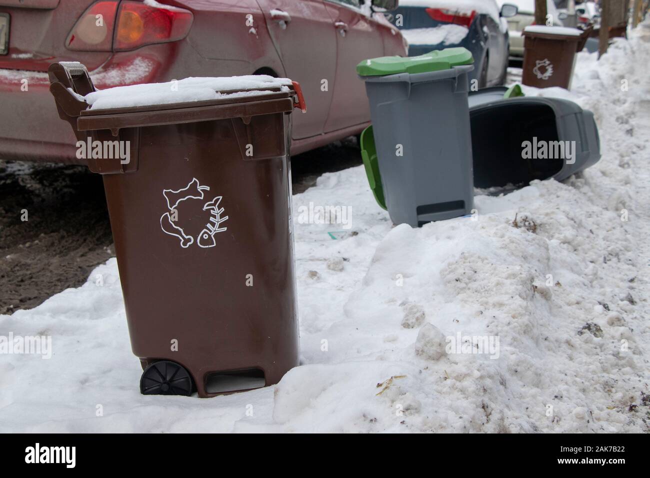 Recycling and compost bins on snowy sidewalk along street and cars in