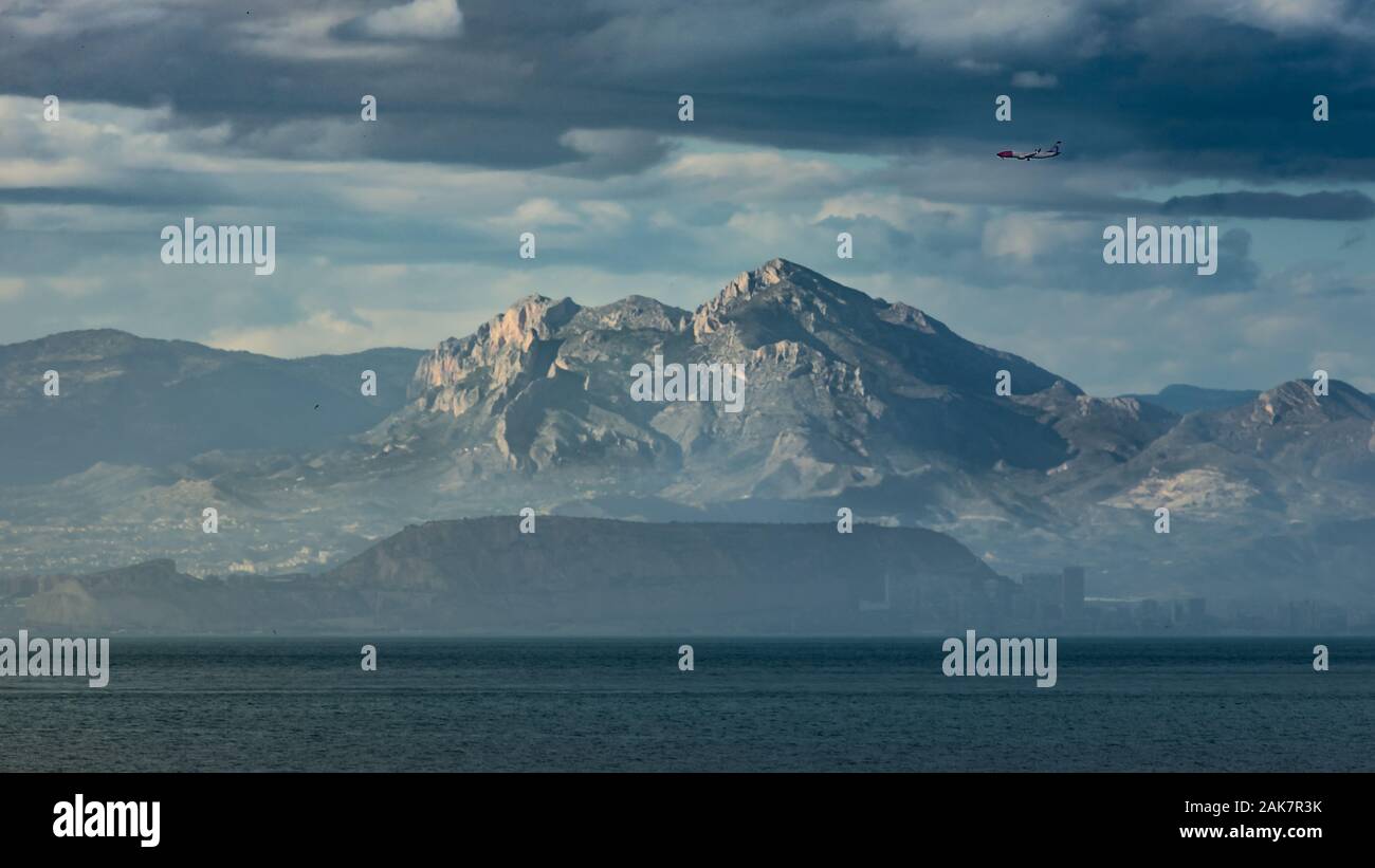 A plane is flying above the mountains in a cloudy sky, a city coast and the sea on the bottom part covered by fog Stock Photo