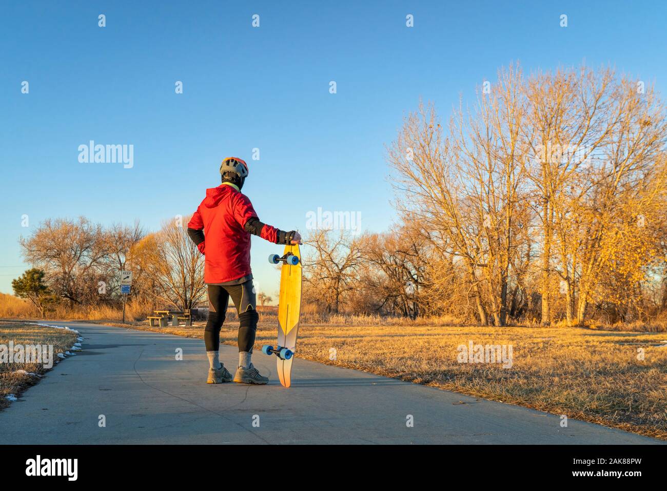 mature man wearing helmet and protective gear is standing with a long skateboard on a paved bike - Boyd Lake State Park in northern Colorado, winter s Stock Photo