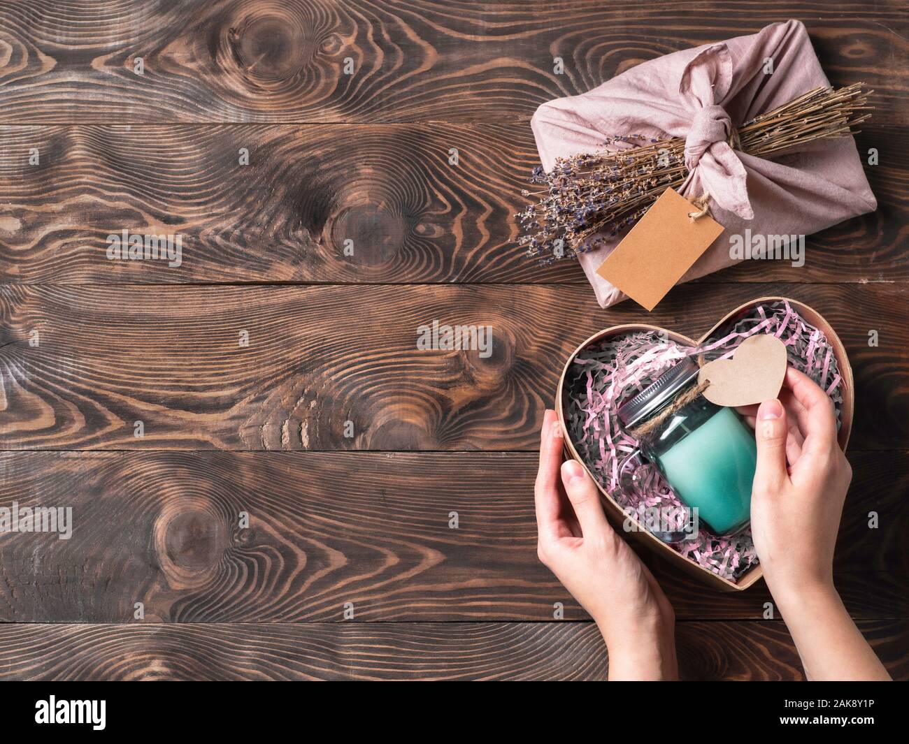Hands hold gift box in heart shape with candle in glass jar for zero waste Valentine's Day. Top down view or flat lay. Copy space for your greetings card, text or design. Brown wooden background. Stock Photo