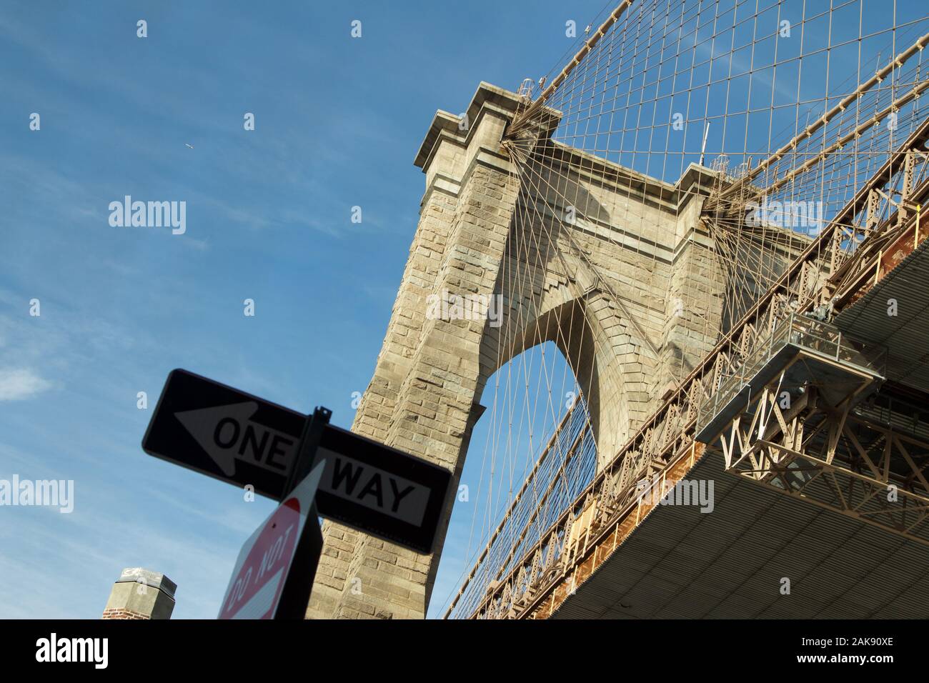 Iconic image of Brooklyn Bridge looking up on a blue day with one way street sign in foreground. Stone suspension bridge with wires seen from low look Stock Photo