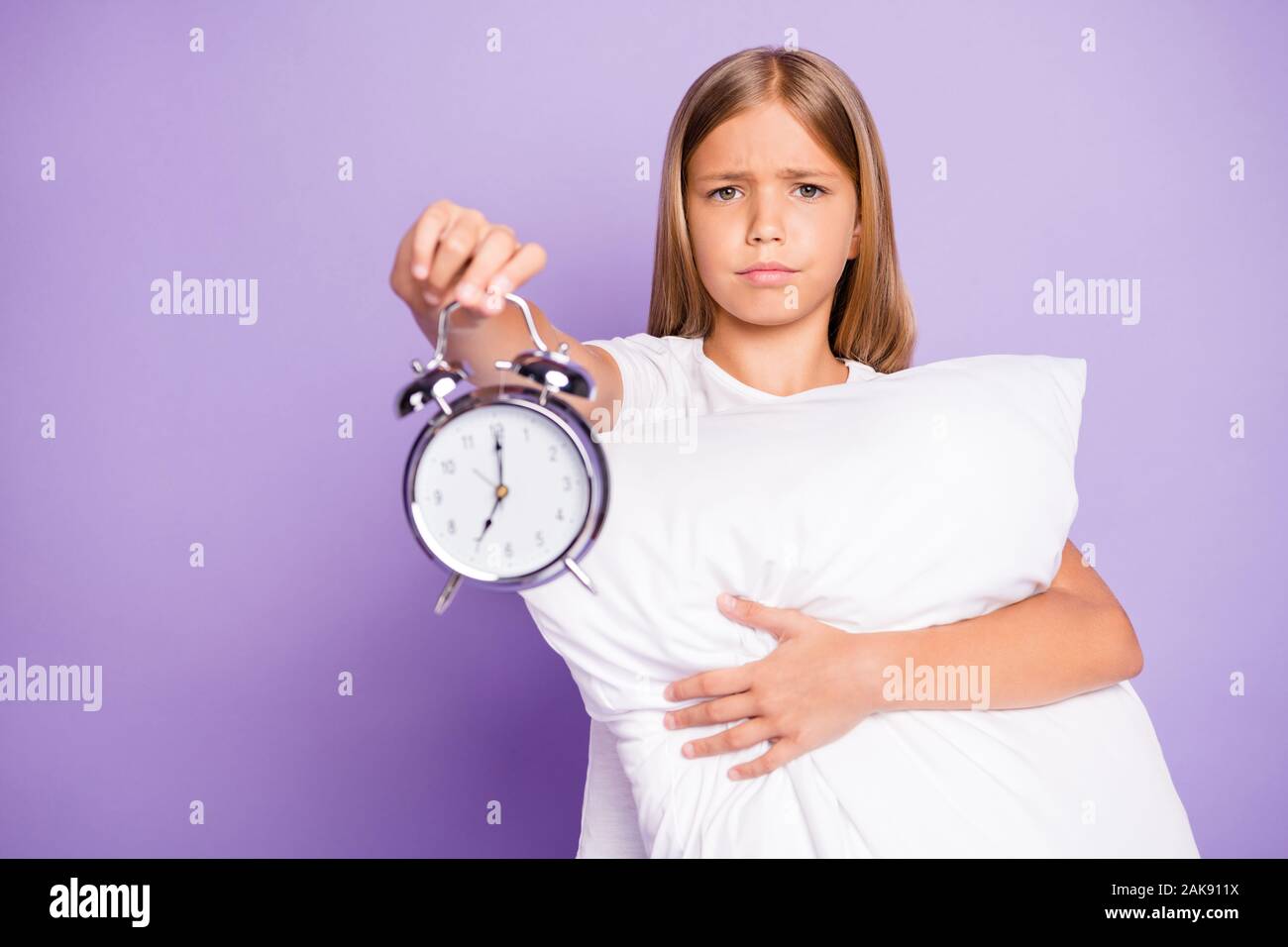 Oh 7 oclock again. Portrait of upset disappointed beautiful kid hug hold hand pillow show clock early time dont want awake and go study wear white t Stock Photo