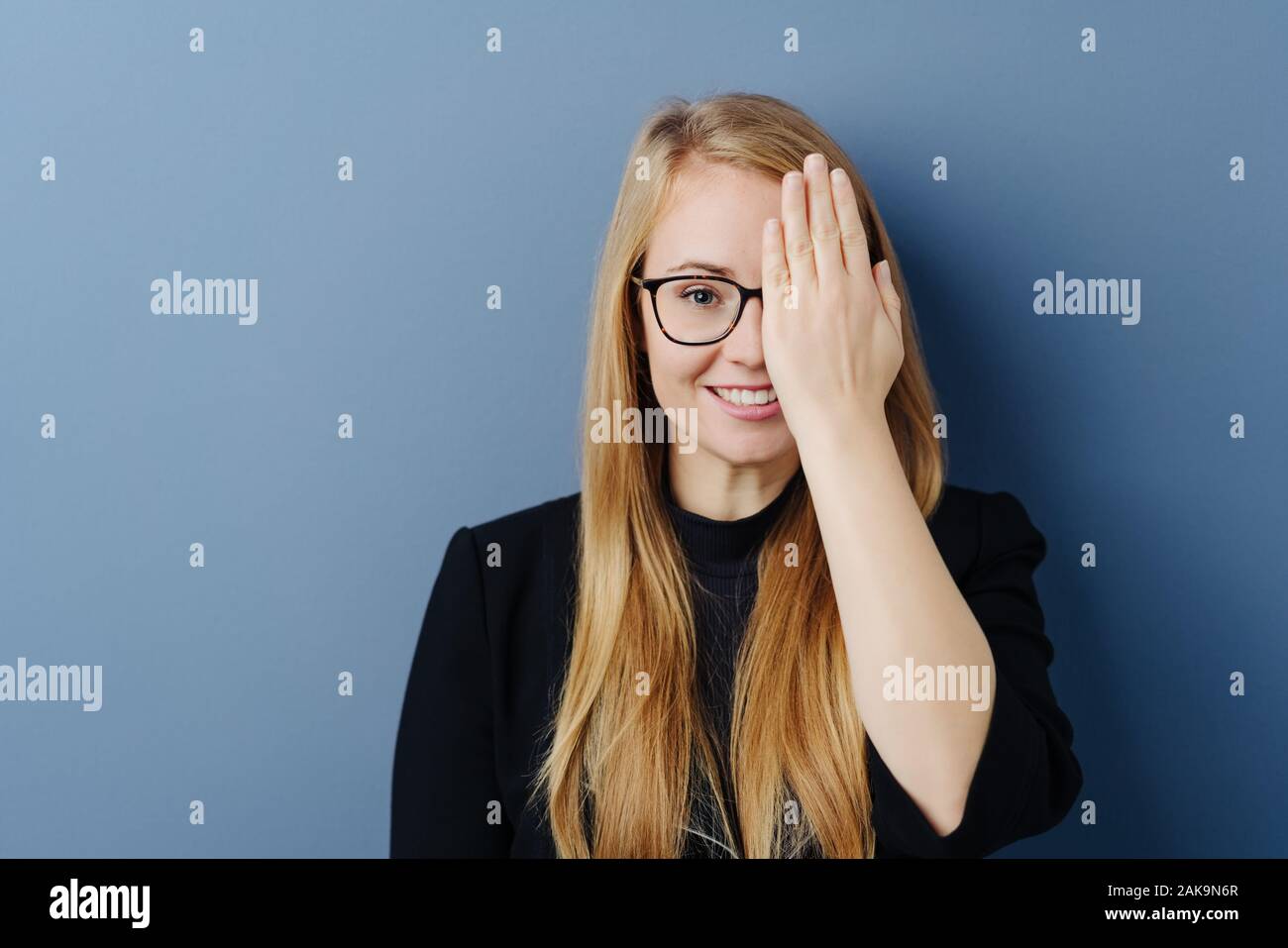 Happy young woman wearing eyeglasses covering one eye with the palm of her hand as she grins at the camera over a blue studio background with copy spa Stock Photo