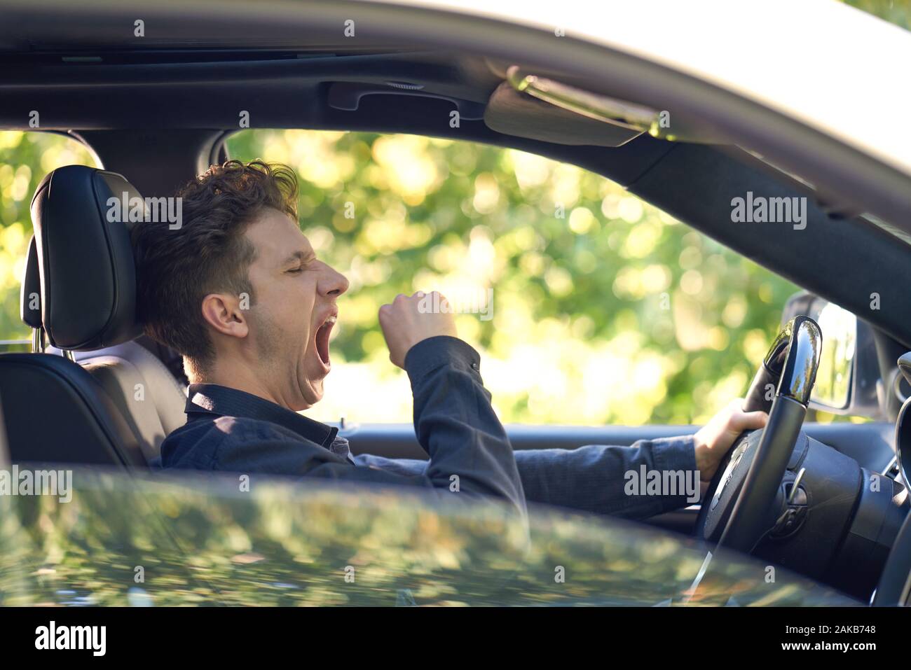 Sideview of a young driver yawning in his car, greenery in background Stock Photo