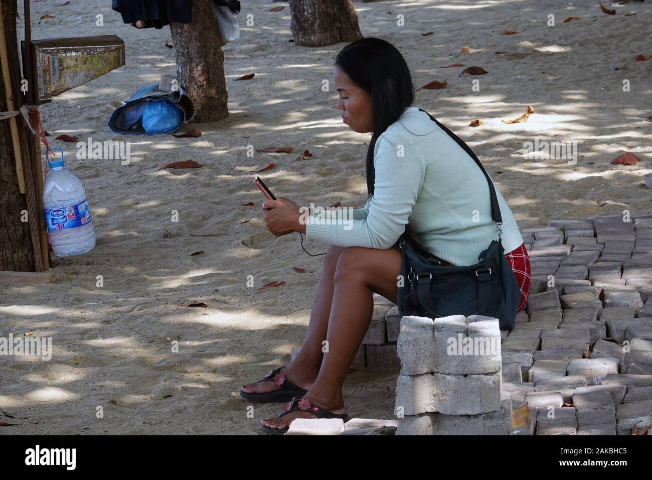 Pattaya, Thailand - December 24, 2019: Woman sitting on pavement tiles near beach and watching her phone. Stock Photo