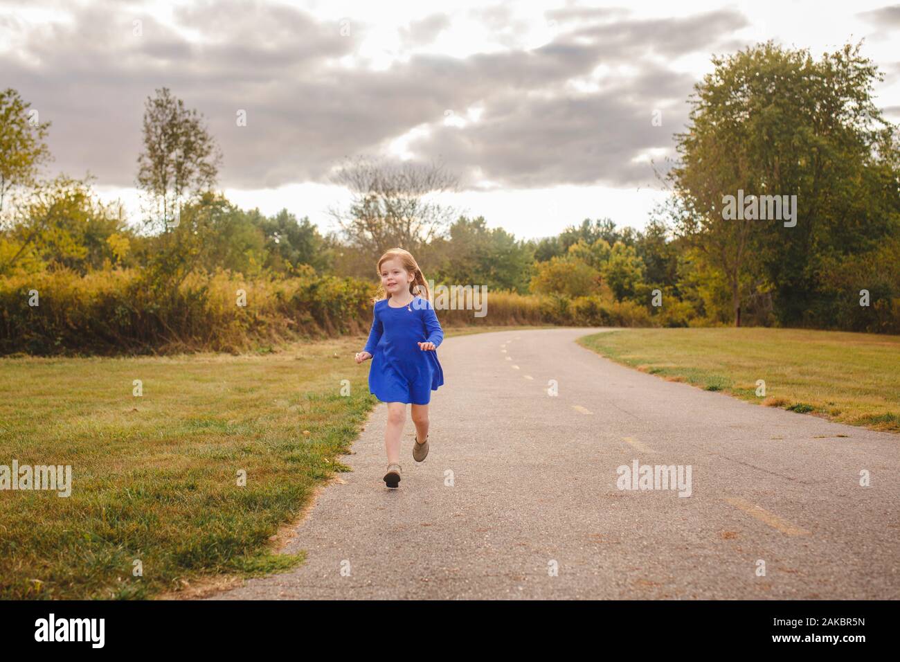 A happy little girl in a bright blue dress runs along a path in a park ...