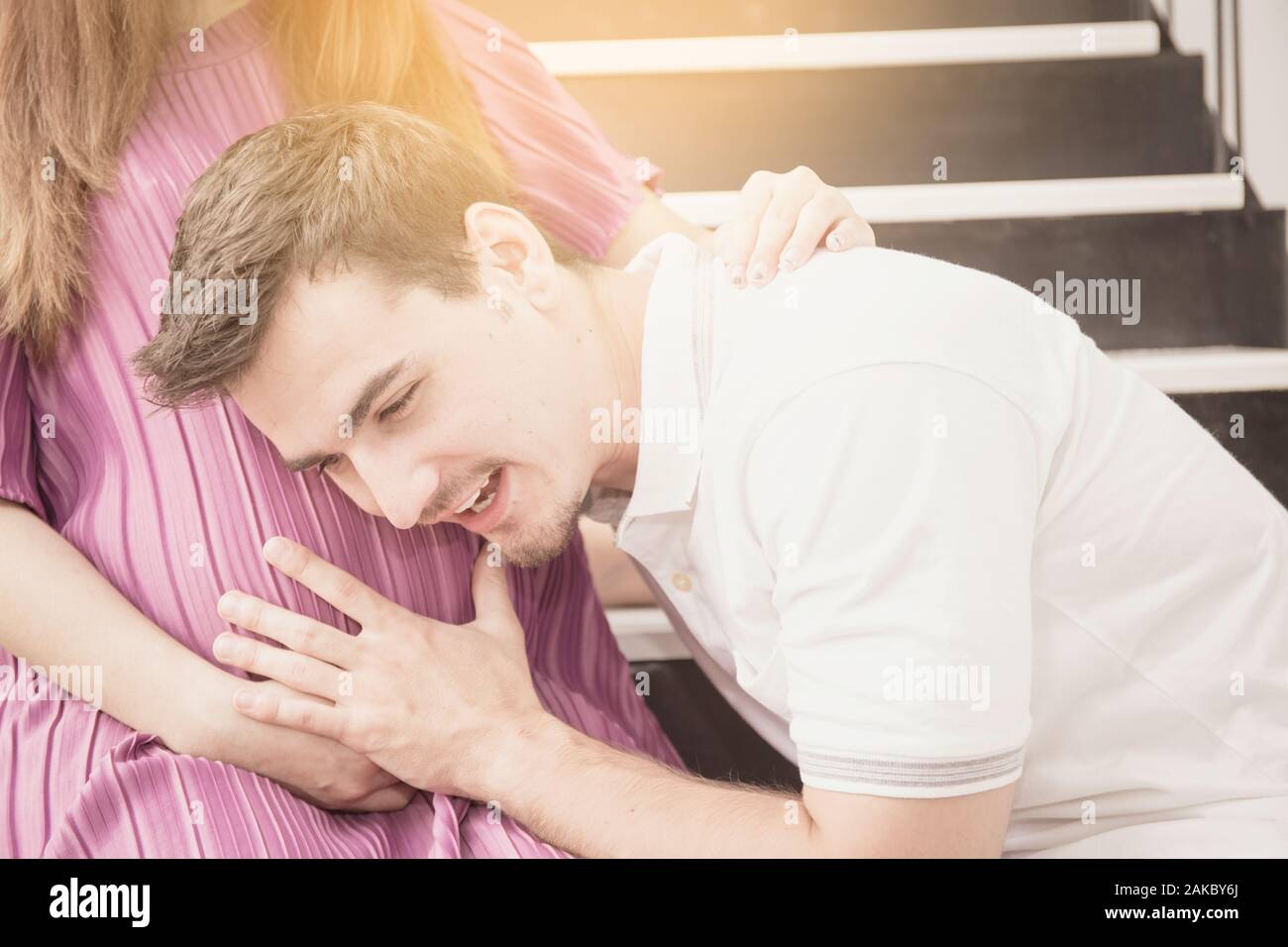 Handsome Asian man and his beautiful pregnant wife are hugging and smiling while standing near the window at home Stock Photo