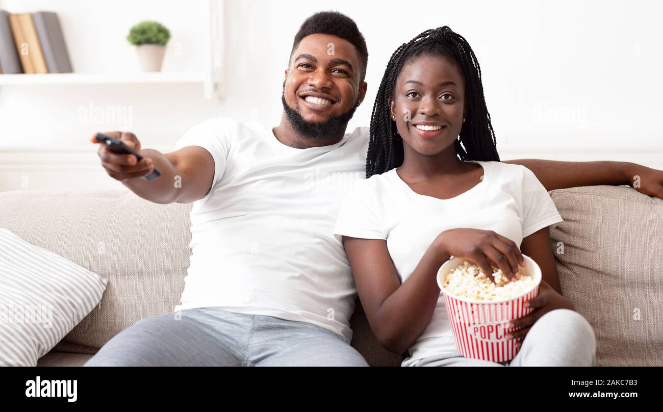 Happy afro couple watching tv and eating popcorn, relaxing on sofa at ...