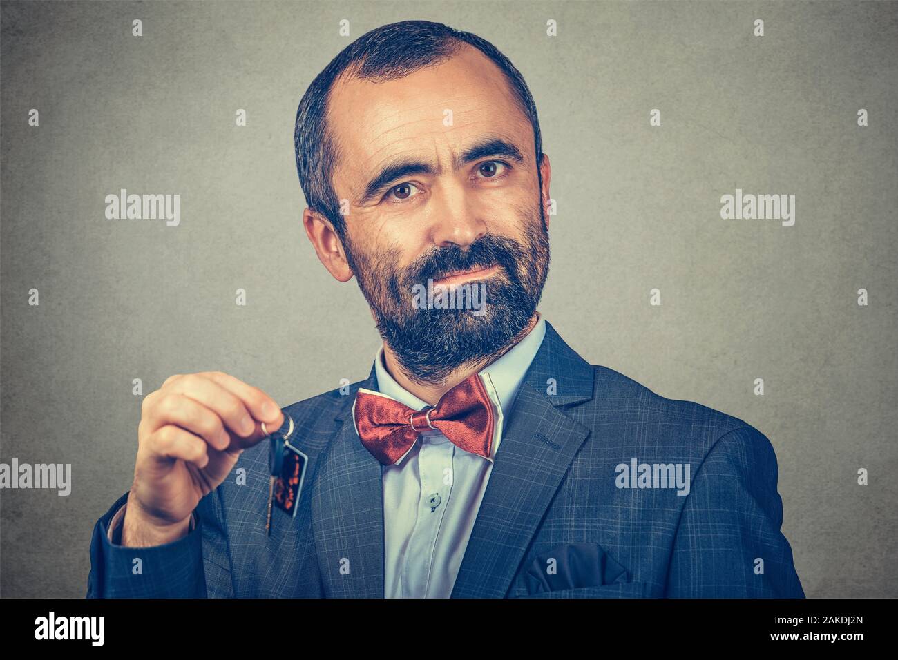 Portrait of serios bearded man, wearing elegant jacket with red bow tie and holding keys from home car office isolated on gray wall background. Human Stock Photo
