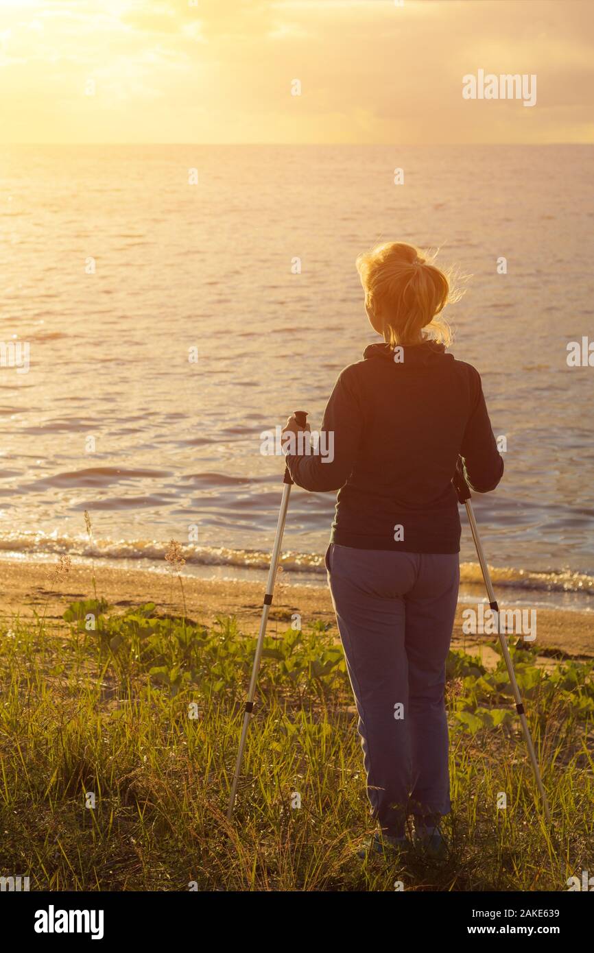 Beautiful young girl blonde in a dark sweater with hiking sticks looks at the sunset. Back view Stock Photo