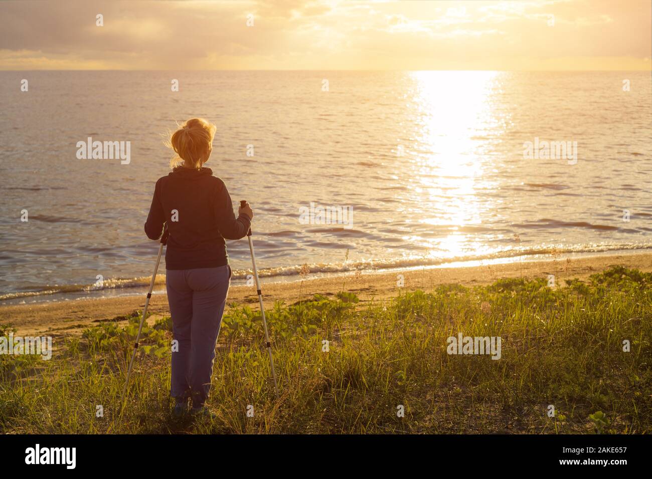 Beautiful young girl blonde in a dark sweater with hiking sticks looks at the sunset. Back view Stock Photo