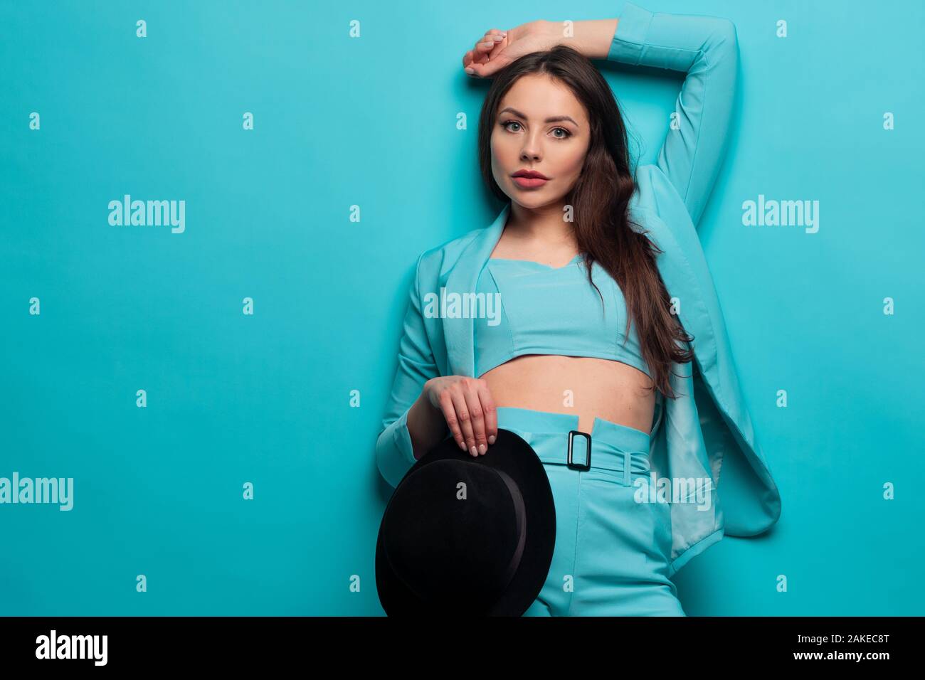 Beautiful and positive brunette in blue on a blue background. Stylish bright image in shades of turquoise. Girl in a jacket and top holding the hat. Stock Photo