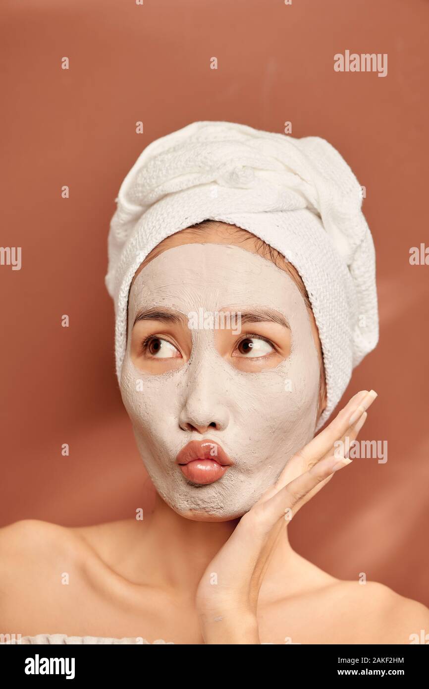 Spa girl with pleased facial expression, applies clay mask on face, gets beauty treatments, wears white soft towel on head Stock Photo