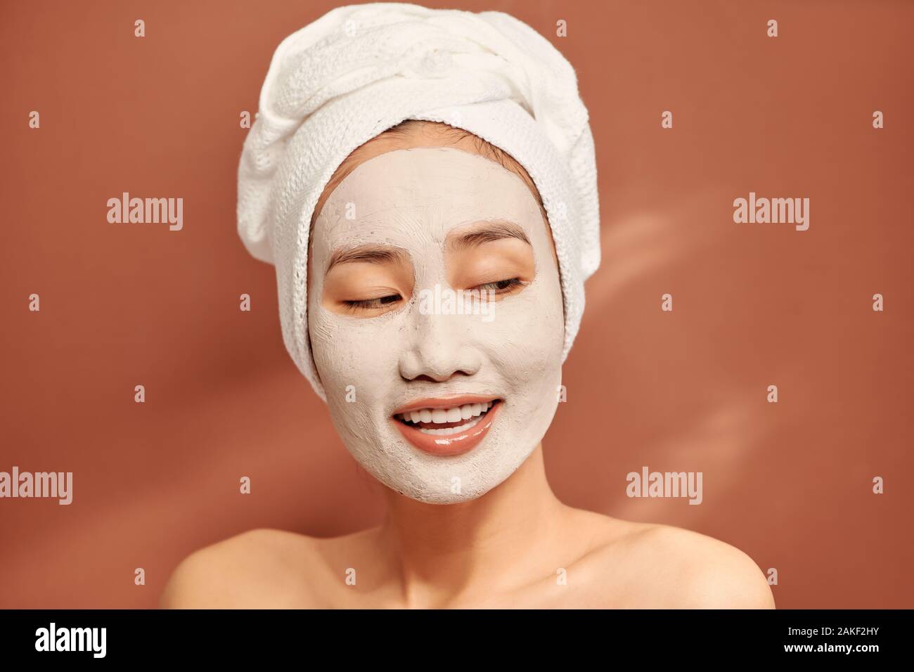 Spa girl with pleased facial expression, applies clay mask on face, gets beauty treatments, wears white soft towel on head Stock Photo