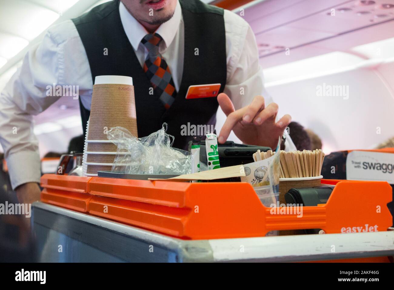 Cabin crew / air steward serves drinks and snacks to passengers from a trolley cart during an Easyjet flight on an Airbus aircraft plane. (115) Stock Photo