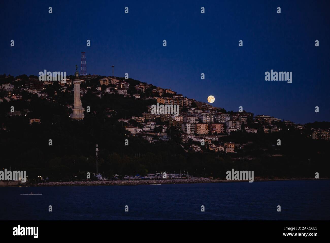 Trieste city skyline view at night with full moon rising from the mediterranean Sea. Famous light tower visible. Stock Photo