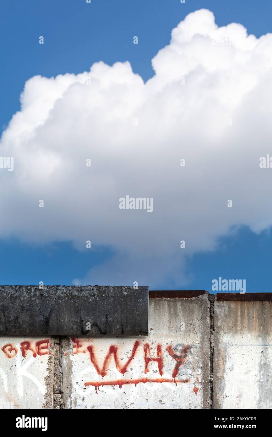 Close up of a remaining portion of the Berlin Wall with a graffiti reading 'WHY', against a summer blue sky with a large puffy cloud Stock Photo