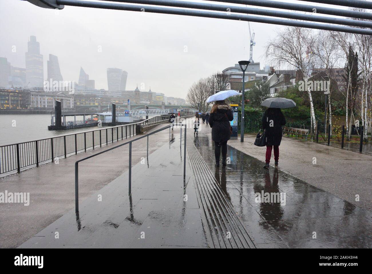 Dark afternoon in wet weather in the city as heavy rain moves in. People with umbrellas walking under Millennium Bridge along South Bank, London, in January Stock Photo