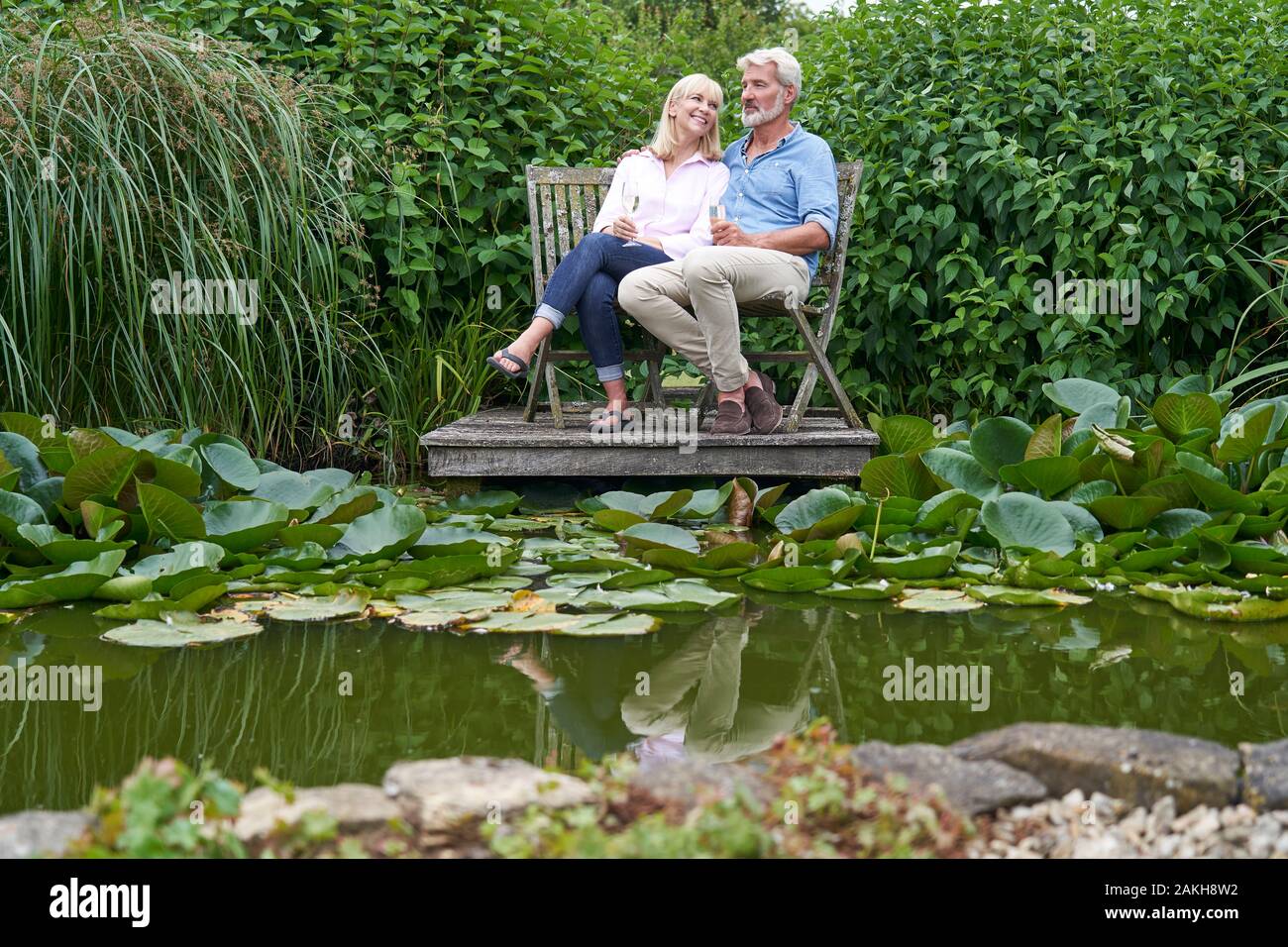 Mature Couple Celebrating With Champagne Sitting On Chairs On Wooden Jetty By Lake Stock Photo