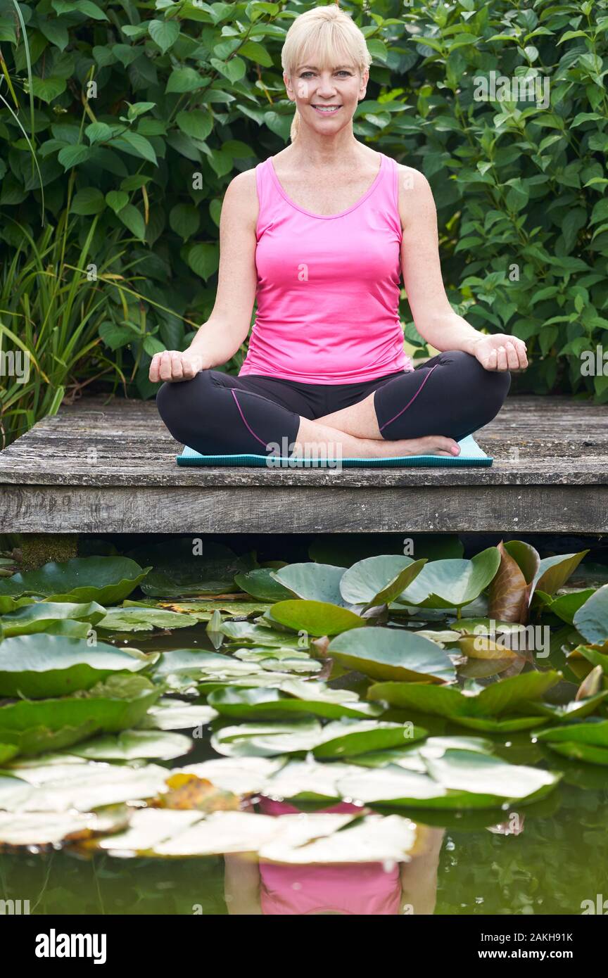 Portrait Of Mature Woman In Yoga Position On Wooden Jetty By Lake Stock Photo