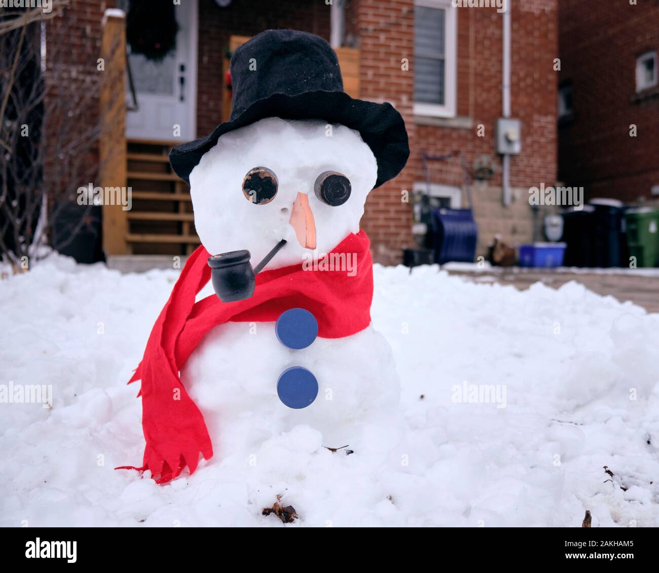 Snowman dressed with red scarf black felt hat blue buttons smoking a pipe in front of Leaside residence Stock Photo