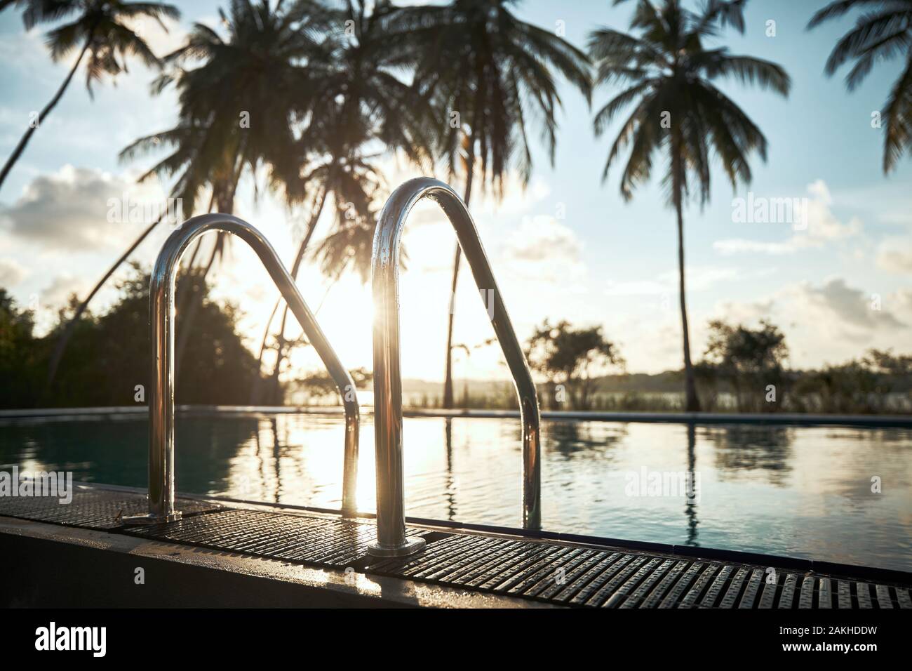 Relaxation in tourist resort. Swimming pool in the middle of coconut palm trees against lagoon at sunset. Stock Photo