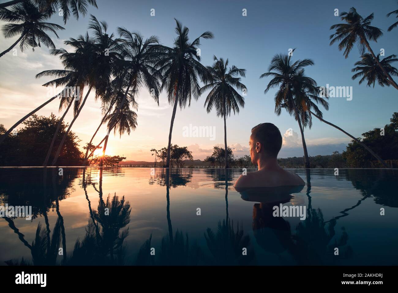 Young man watching sunset from swimming pool in the middle of coconut palm trees. Stock Photo