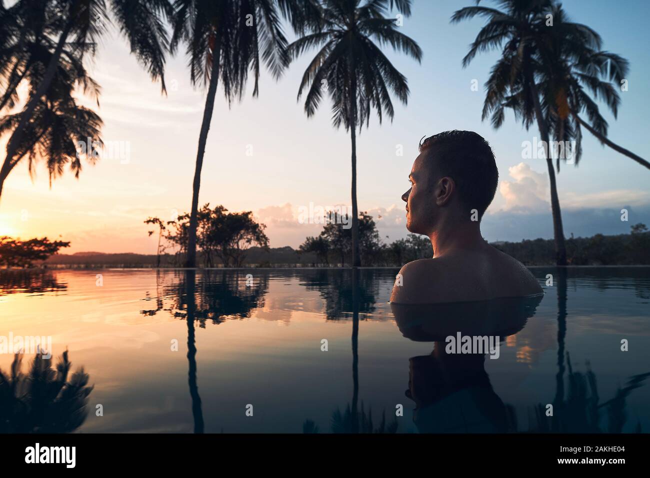 Young man watching sunset from swimming pool in the middle of coconut palm trees. Stock Photo