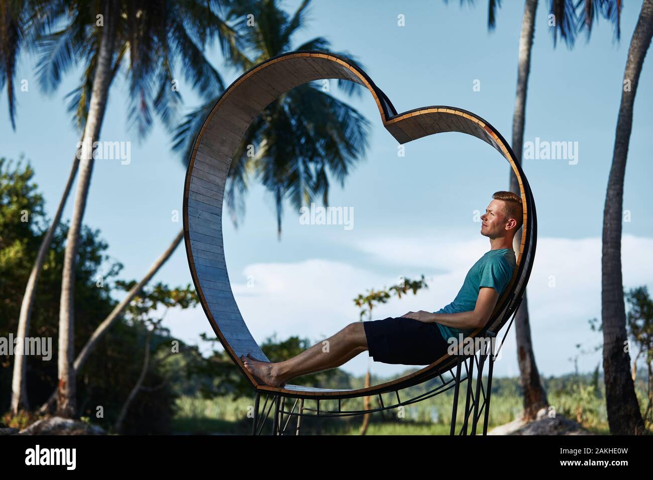 Relaxation in tropical paradise. Young man sitting in wooden heart against palm trees and tropical landscape. Stock Photo