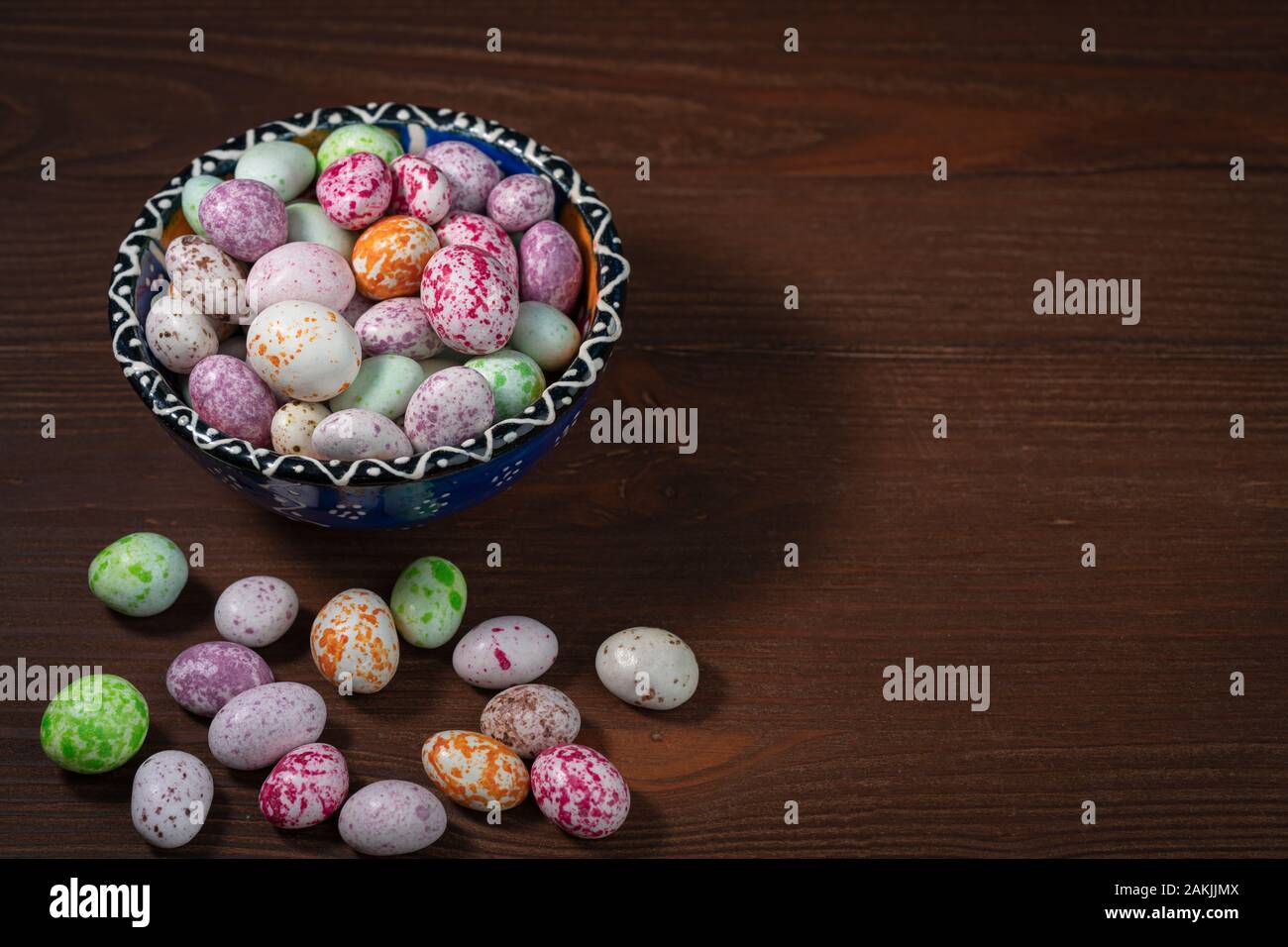 A brightly illuminated blue cup with a pattern stands on a brown wooden background. Inside the cup and nearby are multicolored sweet dragees. Stock Photo