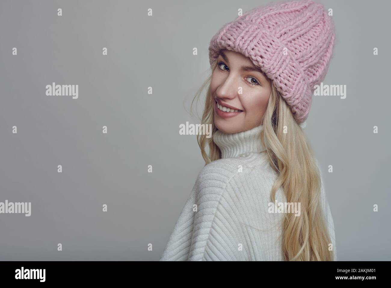 Happy young woman with long blond hair in pink knitted winter hat looking at the camera with a grey background Stock Photo